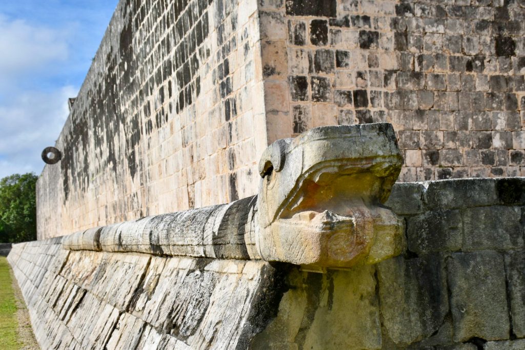 A long stone wall reaches from the left of the frame to the horizon on the left. You can see a stone circle on the wall half way down. Along the bottom of the wall is a stone serpent, whose head faces the camera, mouth open