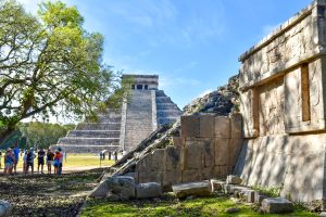 View across the remains of an old stone structure to a large stepped stone pyramid in the background. Tree to the left and group of tourists standing under the tree
