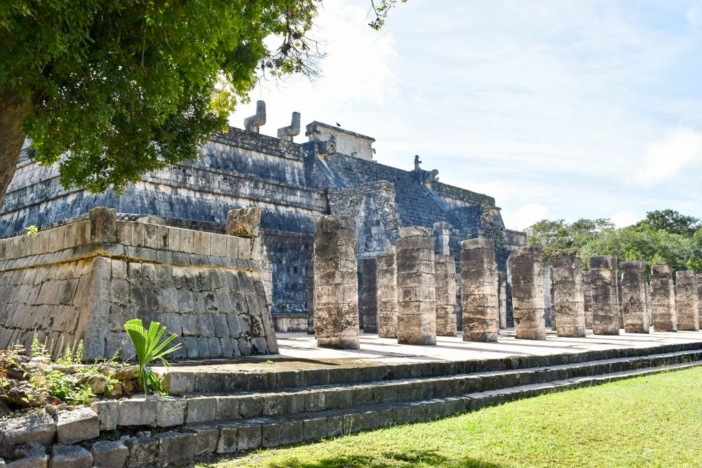 Looking from the left hand side of a wide stone flat topped platform. There is an area at the bottom full of square block columns. Wide staircase to the top of the structure, and some stone figures at the top of the staircase