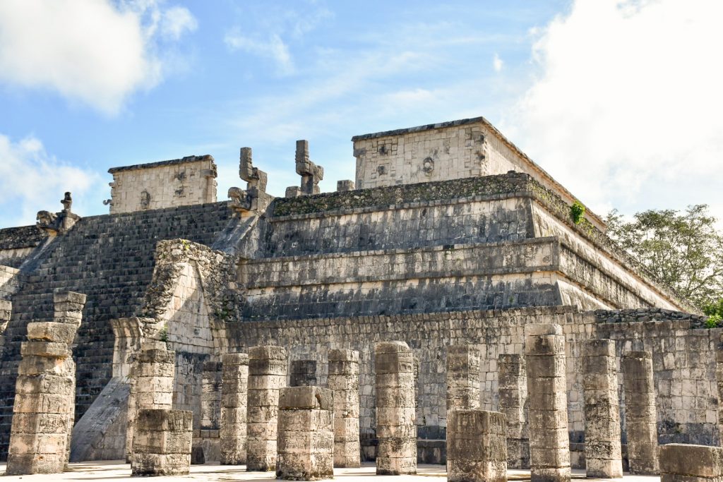 Looking from the right hand side of a wide stone flat topped platform. There is an area at the bottom full of square block columns. Wide staircase to the top of the structure, and some stone figures at the top of the staircase