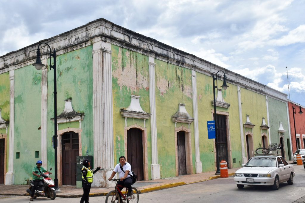 Colourful green and orange buildings on the opposite side of the street. They look weathered and old, with flat roof, white trim and wooden doorways. A white car is driving into the frame from the right and to the bottom left are a man on moped, man on bicycle and traffic policewomen