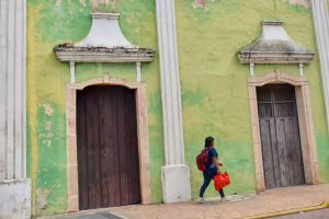 Lady with red backpack and red tote bag walking from left to right of the frame, in front of a lime green wall with two wooden doorways with white hoods over them