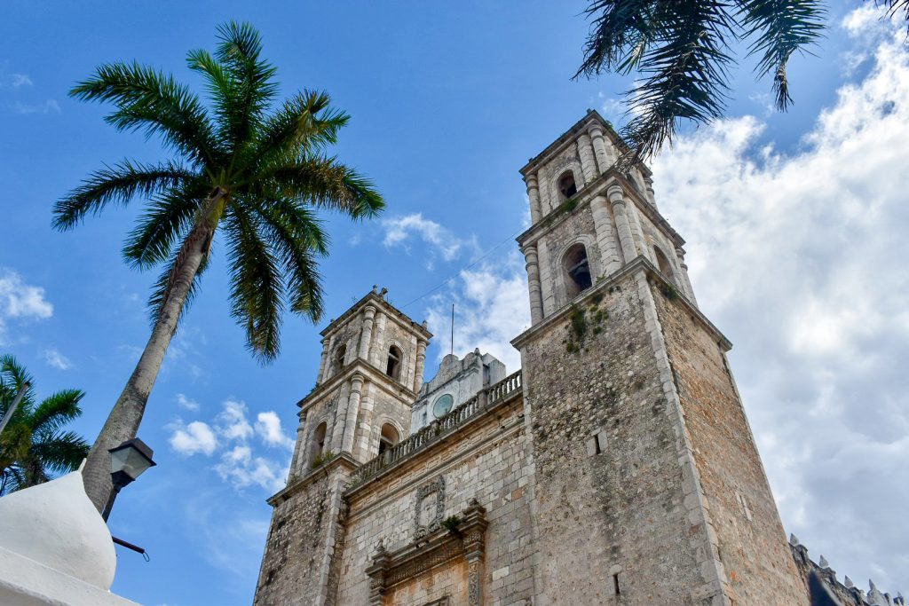 Looking up at the two towers of a Spanish colonial style stone church, palm tree to its left. Blue sky and white clouds