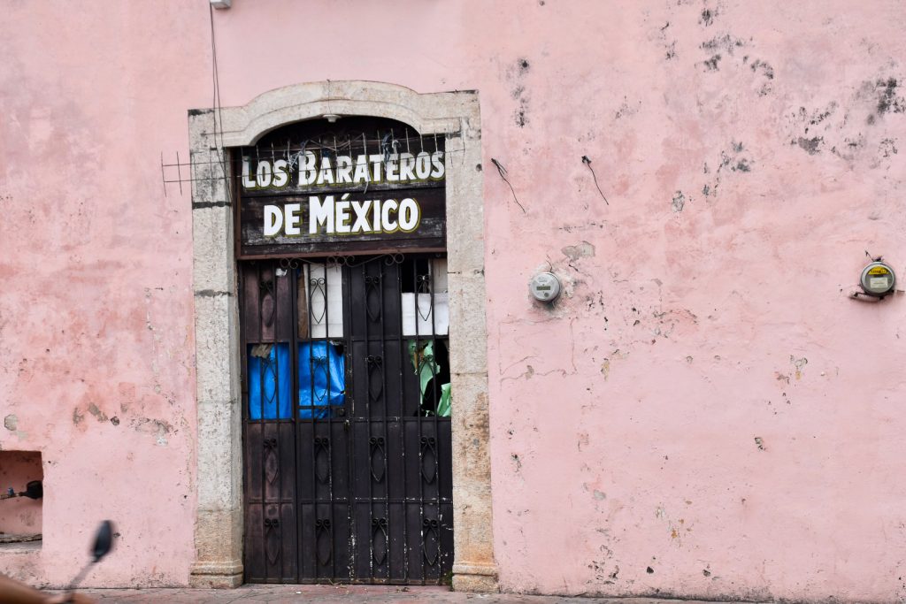 A painted pink wall which looks old and scuffed. To the left of centre is a stone doorway with iron gates and door behind. Above the door it says Los Barters de Mexico