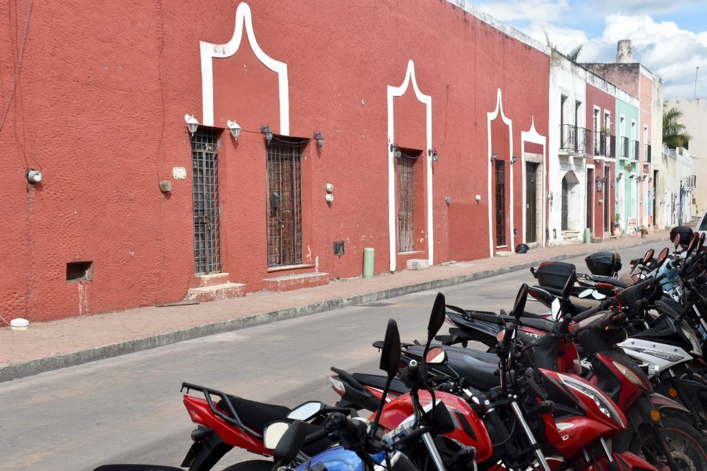 A long building painted rust red sits on the opposite side of the road. In the foreground there is a row of parked motorbikes. Coloured buildings at the very end of the street
