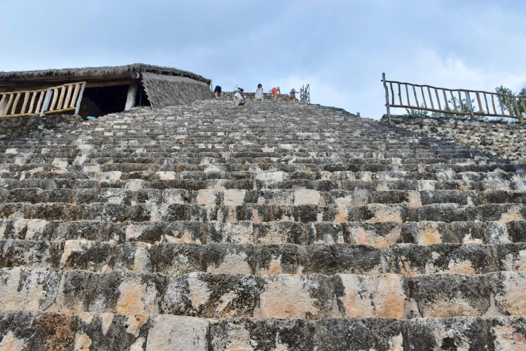 Looking up a tall stone staircase of what might be a temple. There are bamboo handrails to each side around half way up. There are some people making their way to the top of the temple but they look a long way up