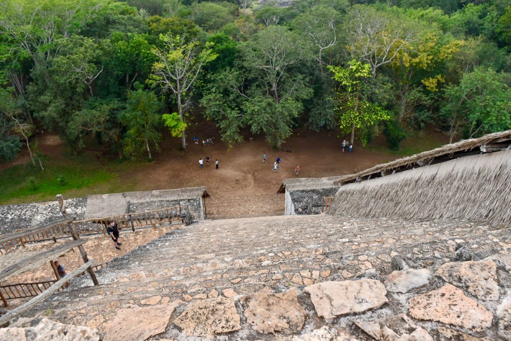 View from the top of what looks like a tall stone building. The height is steep and one person is climbing up the steps but almost on all fours. The people standing at the bottom of the structure look very small. Trees beyond look small as over the tree-line.