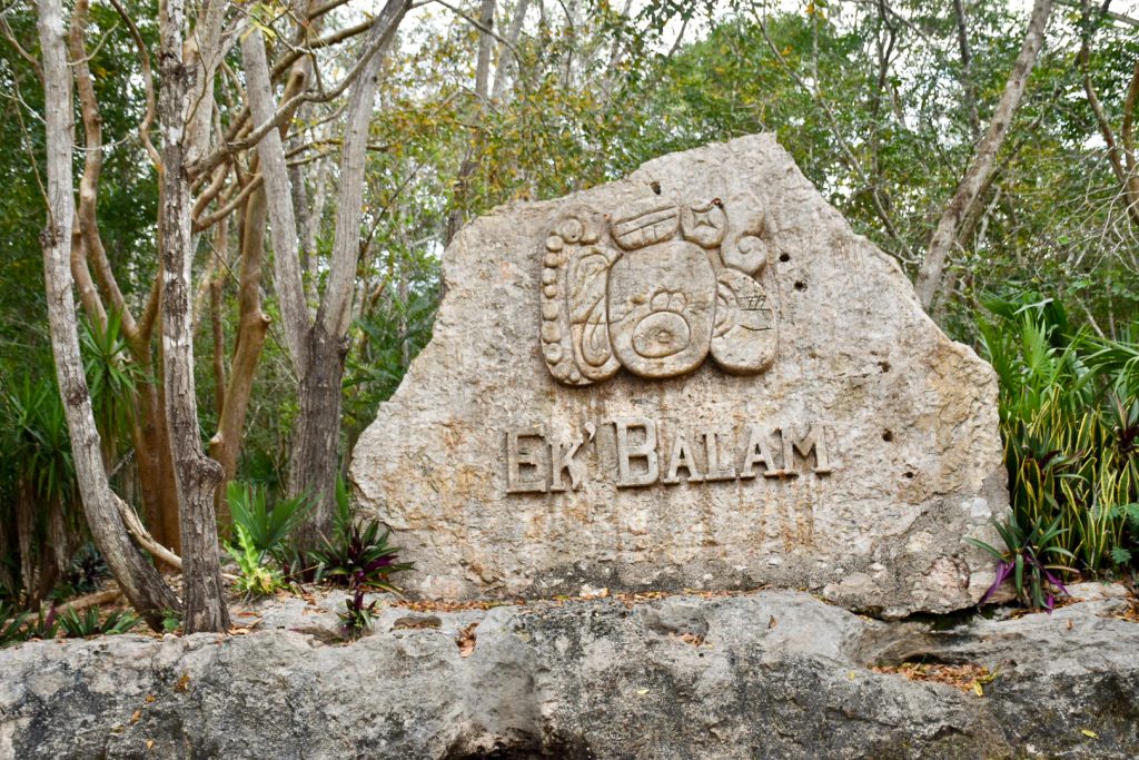 Stone with the words Ek'Balam carved along with a design in the centre of the frame. Stone wall underneath and trees of forest behind