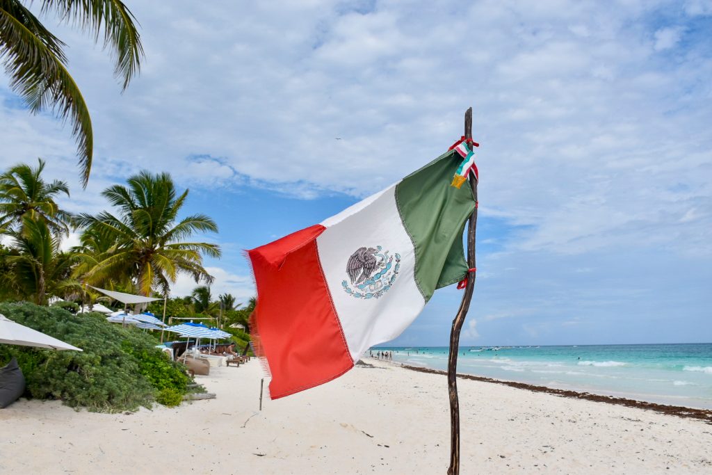 A Mexican flag flies to the left of a wooden post on a white sand beach with palm trees to the left and a turquoise sea to the right. Cloudy blue sky above.