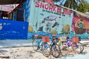 5 bicycles with baskets are parked in front of a building with colourful murals on the side. The most prominent mural is turquoise with a turtle on it and Snorkelling in pink letters, Puerto Morelos in black letters.