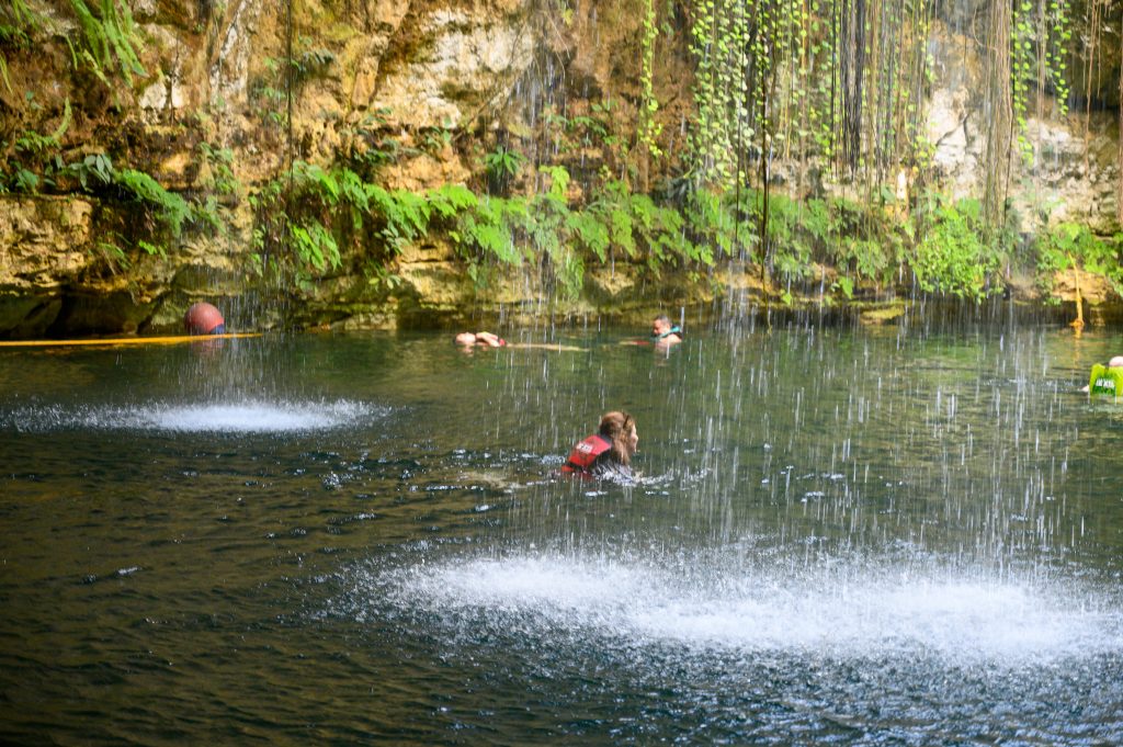 Lady swimming in water with a life jacket on. Water is pouring in from above like a thin waterfall. Rocks and plants in the background
