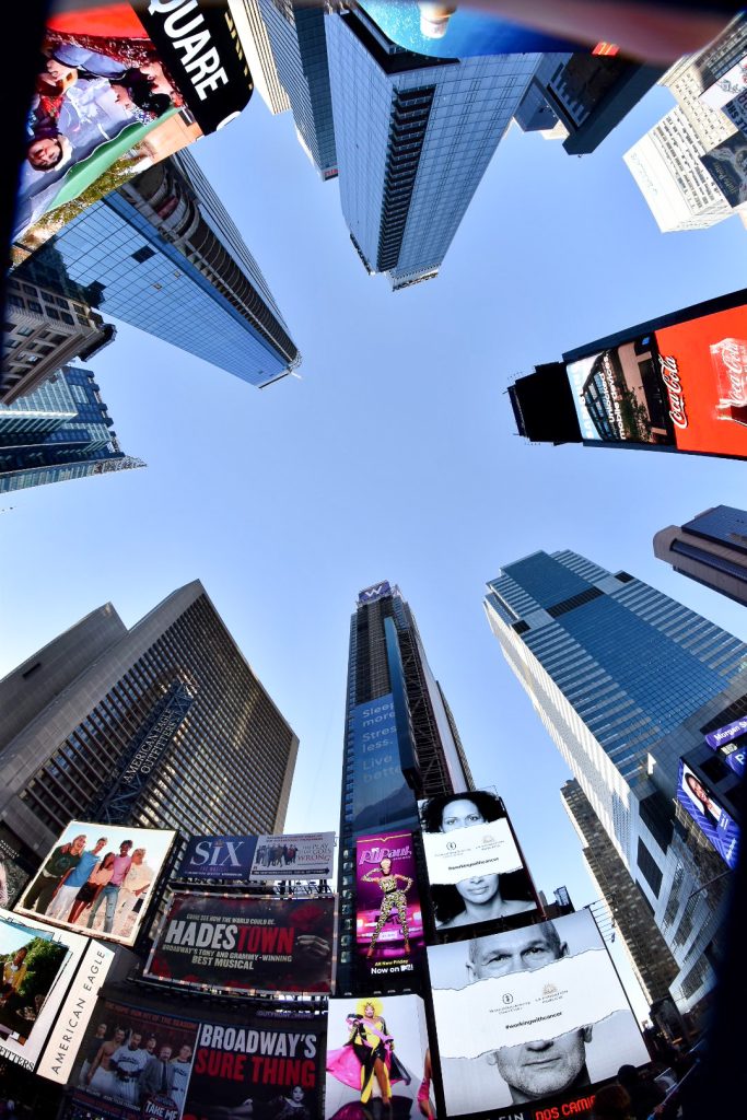 Looking up to the sky which is clear pale blue. Tall skyscrapers around the edge of the photo appear to be leaning into the middle of the photo in a circle. There are blllboards on the bottom buildings advertising Broadway shows