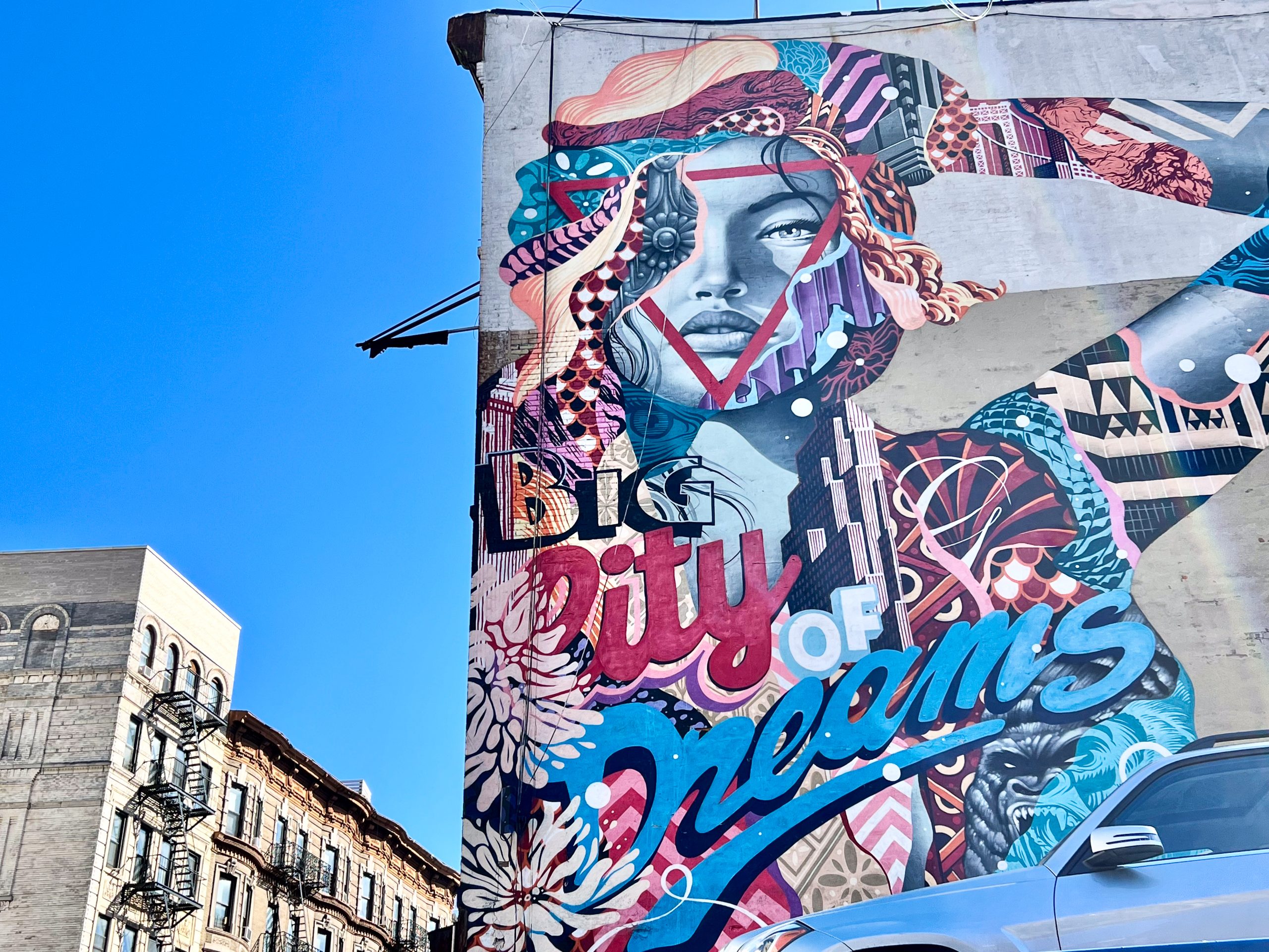 Side of building to the right hand of frame. Features a huge colourful mural of a lady looking at the camera. Features the words Big City of Dreams. Blue sky and top of a building with fire escapes to the left hand side