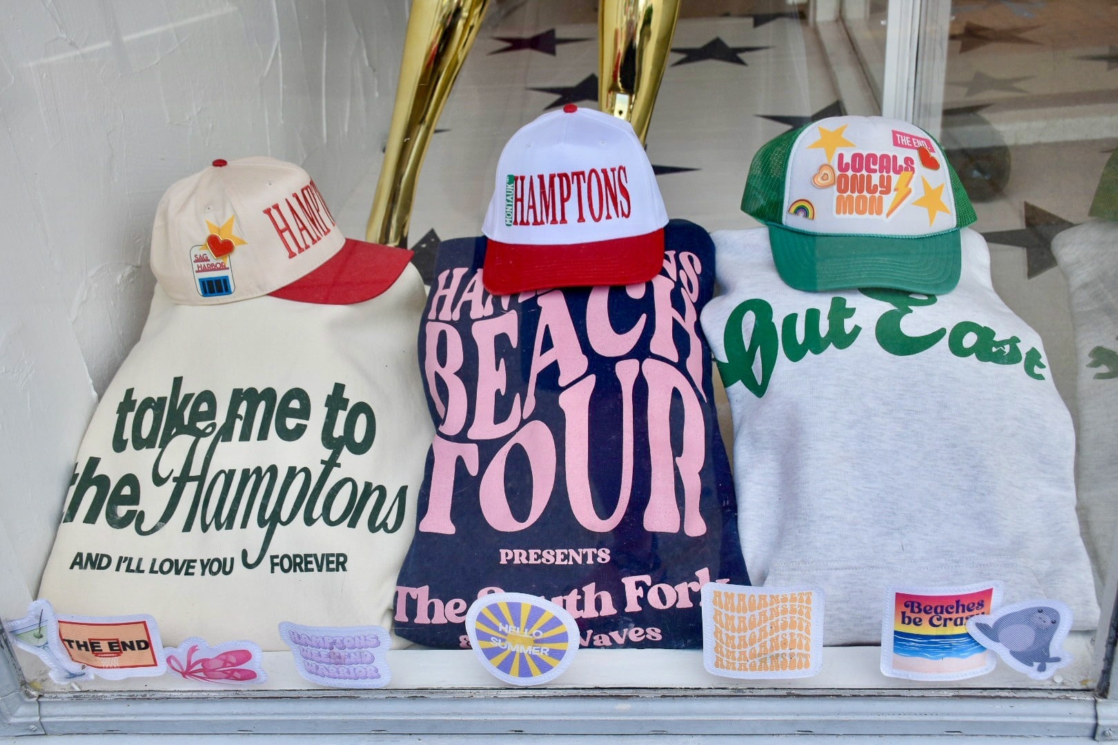 Row of folded sweatshirts in a shop window. From left to right: cream sweatshirt with the logo 'take me to the Hamptons and I'll love you forever', then navy blue with pink writing saying 'Hamptons Beach Tour', then to the right a grey sweatshirt with green writing which says 'Out East'. Each sweatshirt has a Hamptons related baseball cap on top.