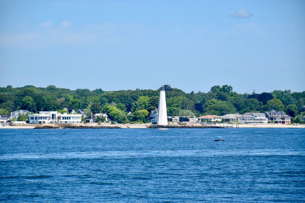 View across a river with a strip of land and trees across the horizon. It has a white lighthouse in the centre and buildings to either side. Pale blue sky above