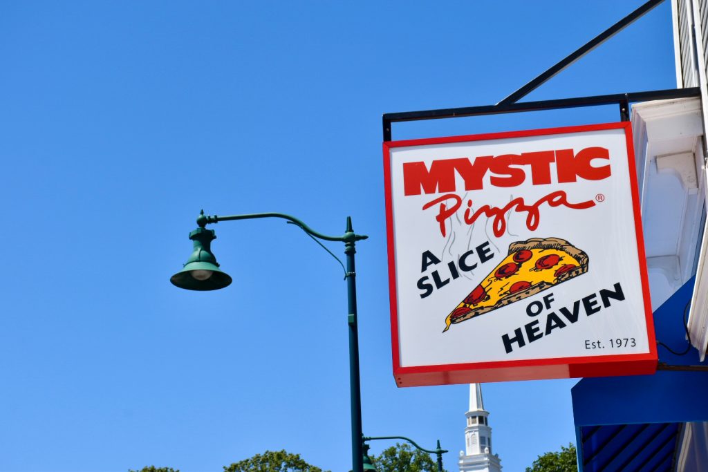 Clear blue sky behind. A square white signpost with a red border hangs from a building to the right. The sign says Mystic Pizza, A Slice of Heaven' on it. Green lamppost in the middle behind the sign.