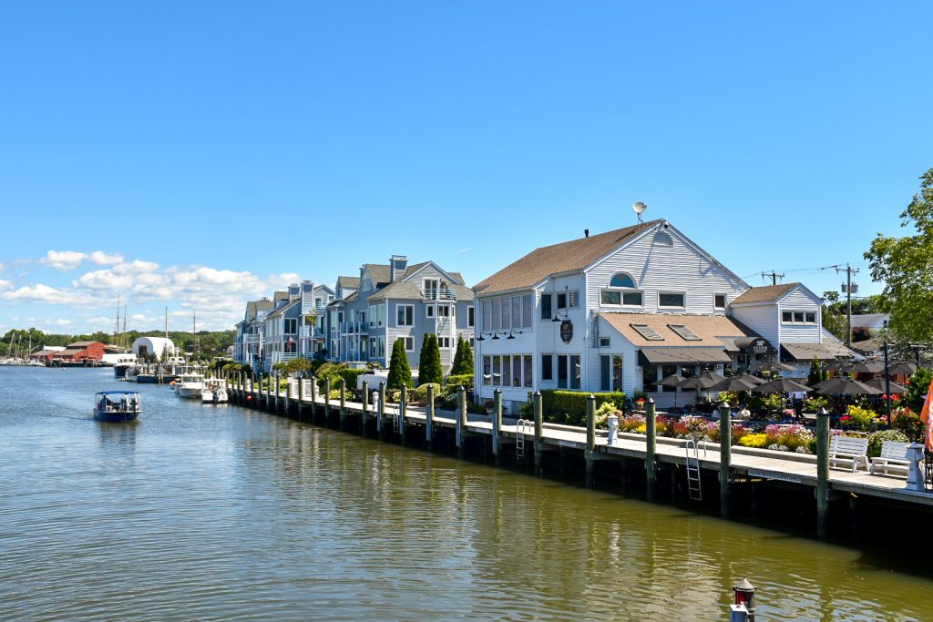 Looking down a river, possibly from a bridge, with a line of large wooden clapboard houses along the quayside to the right. One small boat is making its way down the river towards the camera. Pale blue sky above