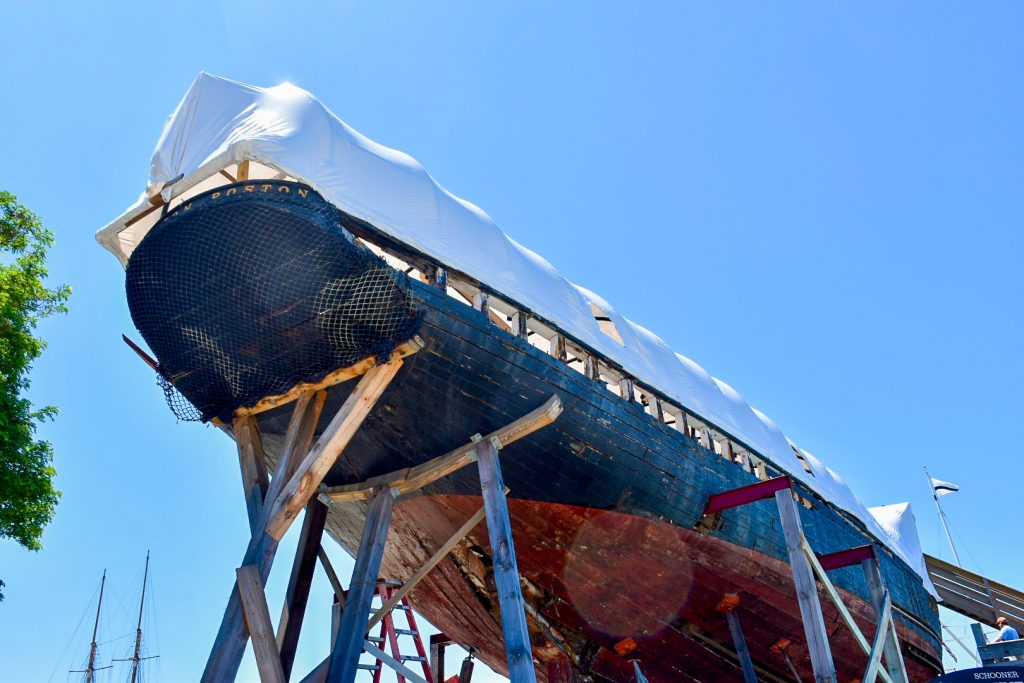 Looking up from the ground to the hull of a wooden ship sitting on large wooden scaffolding. It has a red underbelly and black sound. You can see the word 'Boston' on the back. White sheets over the top. Blue sky behind.