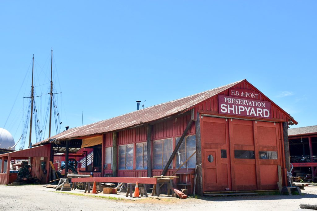 A long red wooden building sits across the centre of the frame with orange cones and workshop items in front. It says 'H.B. DuPont Preservation Shipyard' in white letters on the front of the building. Two tall masts from a sailing ship to the left hand side. Blue sky behind.