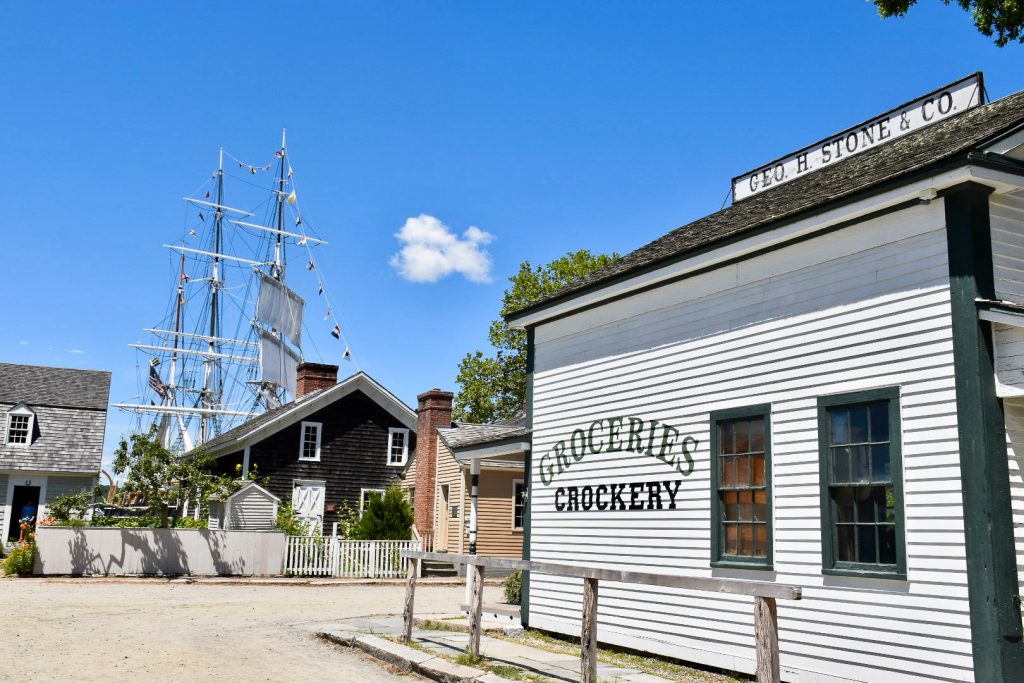View down a short street with a white wooden clapboard building to the right which says the words 'Groceries. Crockeries. Geo H Stone & Co'. Straight ahead at right angles is a row of houses. Tall masts of a sailing ship can be seen behind. Blue sky beyond.