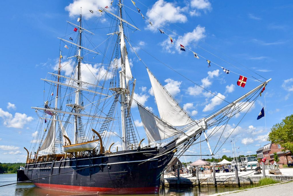 A large sailing ship or schooner sits in the bottom of the frame with a black hull and red strip just above the water. The three tall masts of the ship fill the rest of the frame. They are connected by rigging and ropes. There is a row of shipping flags at the front of the ship.
