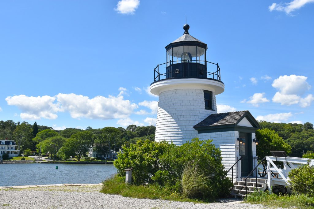 A small wide white lighthouse sits on the edge of a river with trees on the far side. Blue sky above. The lighthouse has a black painted light tower at the top, and it has a vaulted doorway to the right hand side