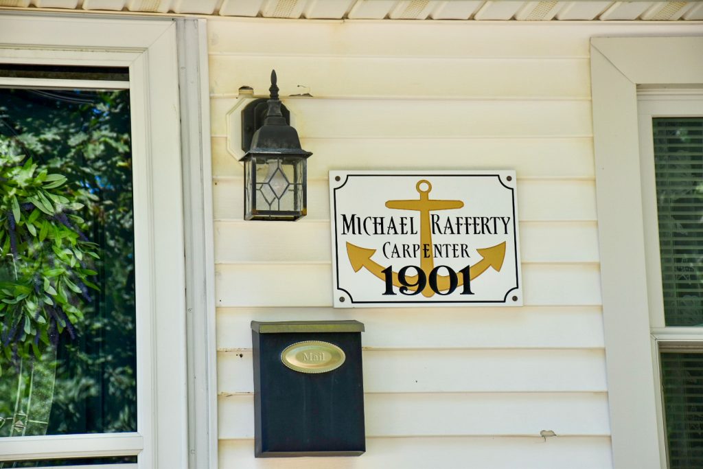 White wooden clapboard building with a doorway to the left and window to the right. In the centre is a black lantern light on the wall, a black mailbox and a white sign saying 'Michael Rafferty, Carpenter, 1901'
