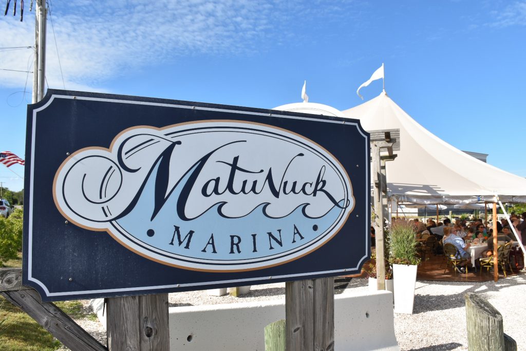 Navy blue sign boat in bottom left segment of frame. It has a white oval with the words 'Matunuck Marina' on it in navy above pale blue wave design. A large white marquee behind. Pale blue sky above