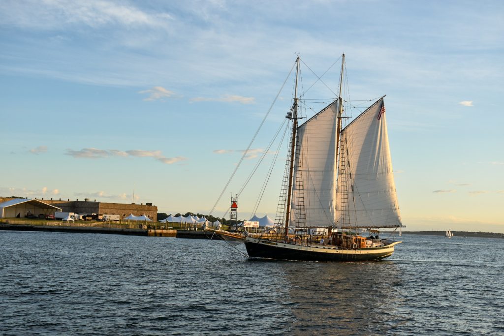Image of an elegant sailing boat with two white sails out heading to the left across a harbour. Low rise buildings on the horizon. Pale blue sky above