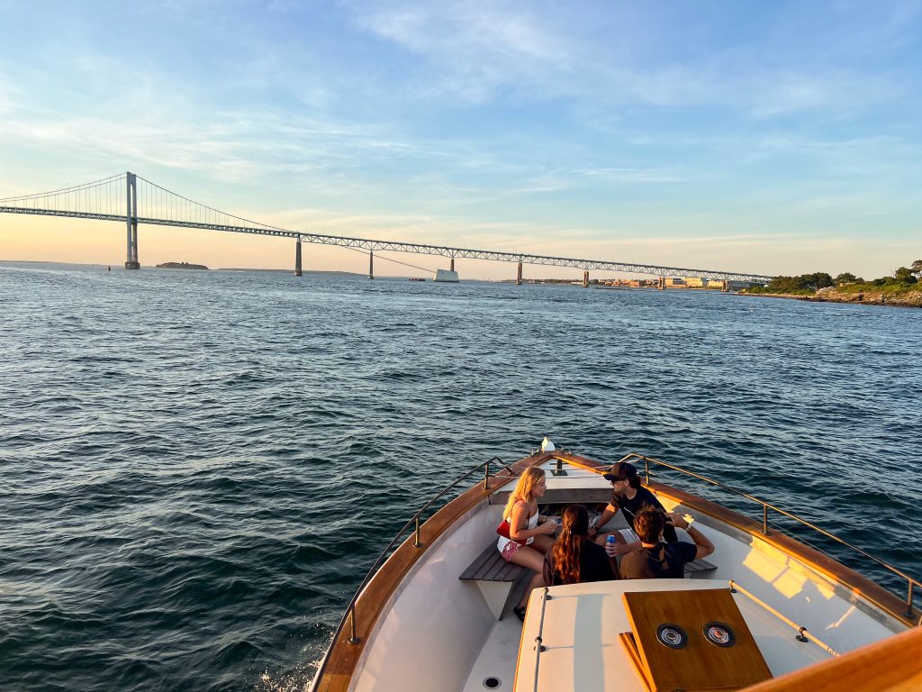 A white boat with tan wooden trim is sailing out to harbour towards a suspension bridge on the horizon. 4 passengers are chatting at the front of the boat in the light of golden hour