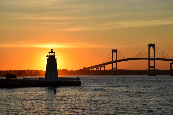 View across a harbour at sunset with a two span suspension bridge silhouetted on the horizon. Into the left is a lighthouse in silhouette with the sunset directly behind it.