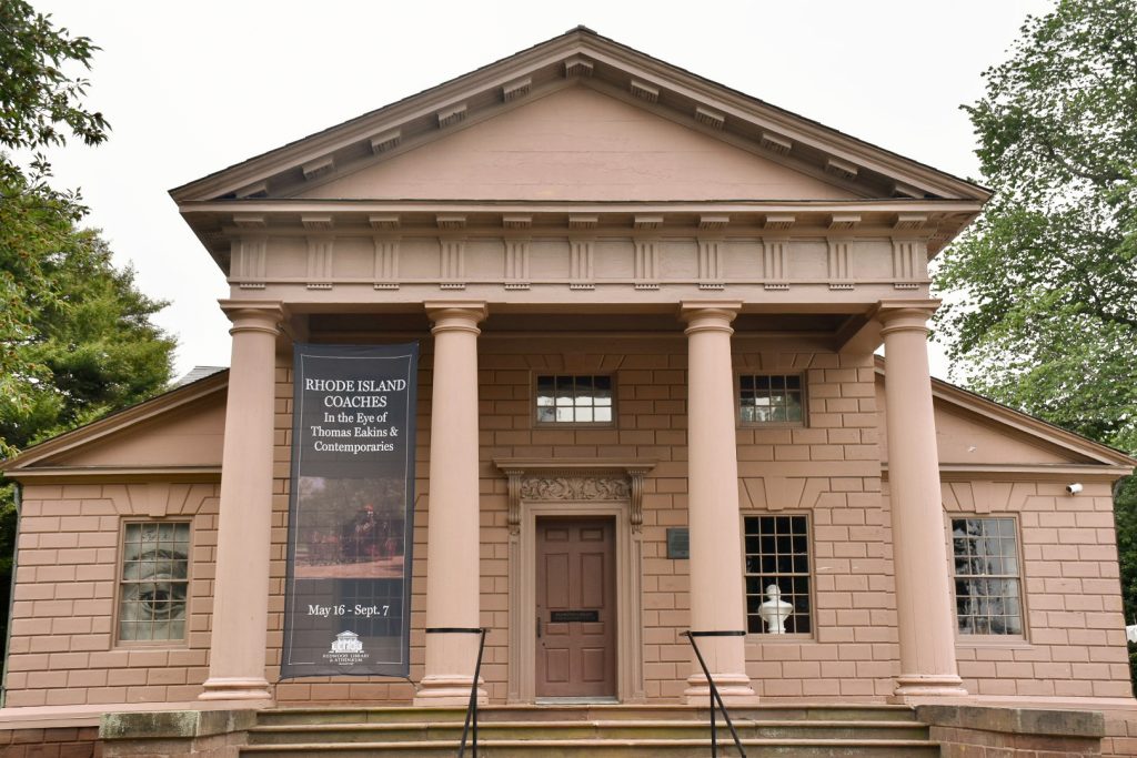 A large detached building faces the camera with four Greek revival style columns in front beneath a triangular roof. Symmetrical design with central door and two windows either side. Painted salmon pink.