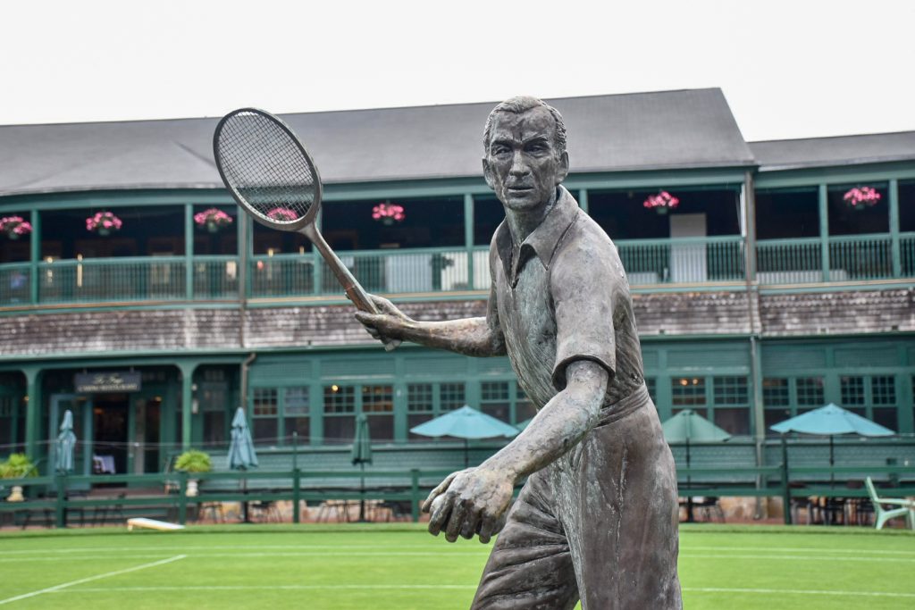 A statue of a male tennis player is facing the camera with a two storey green pavilion building and grass tennis court behind. The statue is holding a racquet in right hand, raised as if to receive the ball