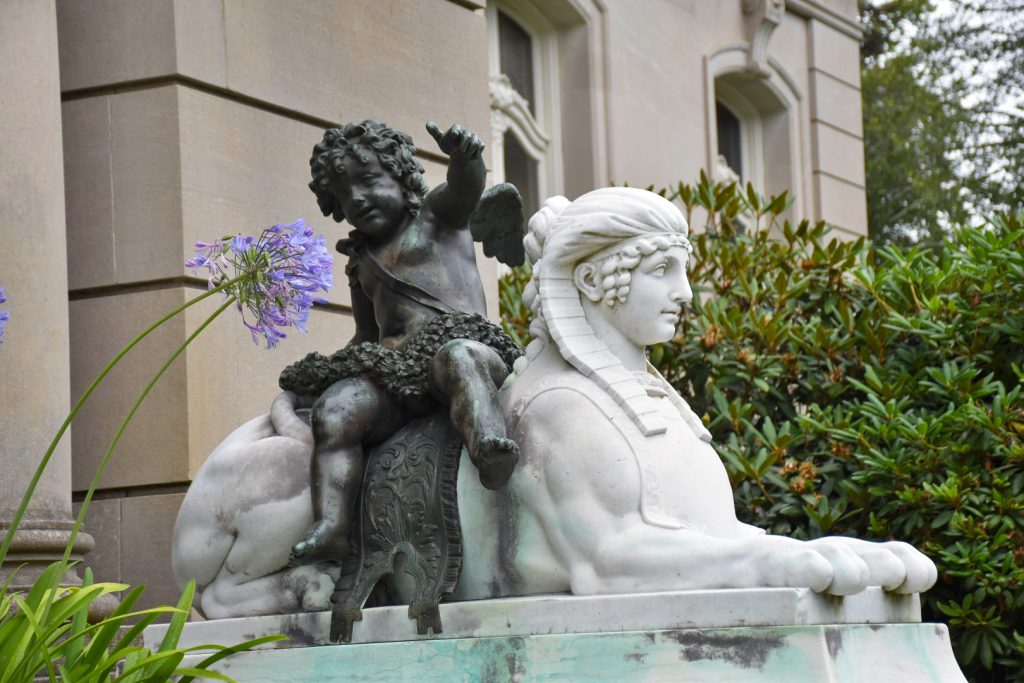 A white marble carved sphinx sits outside a stone building. It has a lady's face and has a brass statue of a cheeky male toddler sitting on its back