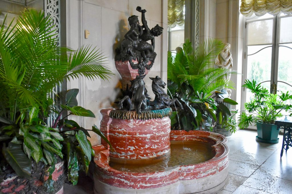 Inside a conservatory filled with palms either side of a stone water fountain with black statues of horses and water nymphs