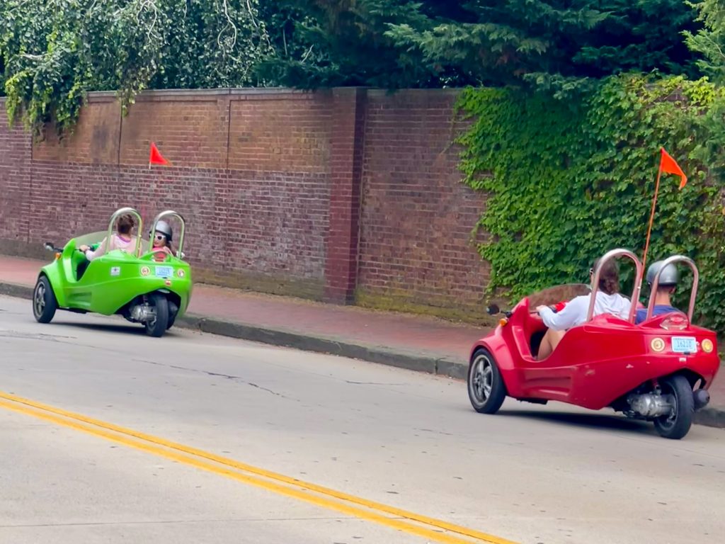 Two small three wheeler cars are making their way across the frame on a road from right to left. Red car to the right and green car to the left. Brick wall behind with grass above.