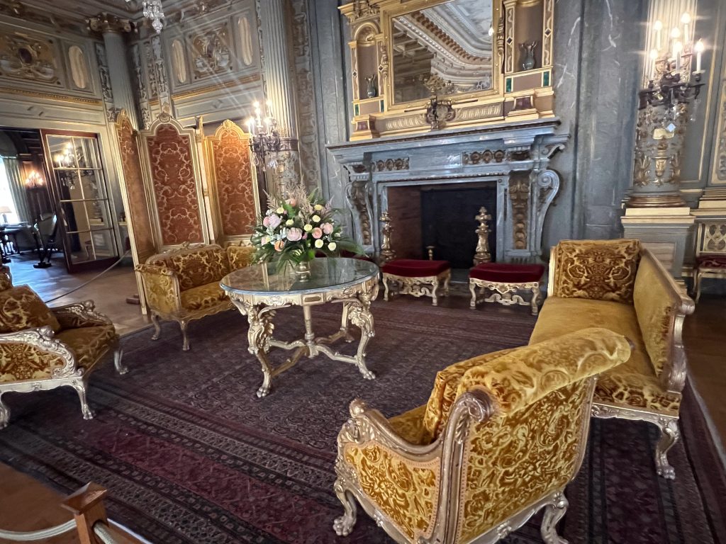 Old fashioned chairs with ornate gold legs and gold velvet upholstery sit around a glass and hold table in the centre of a marble room. Very grand. Large burgundy and navy carpet beneath