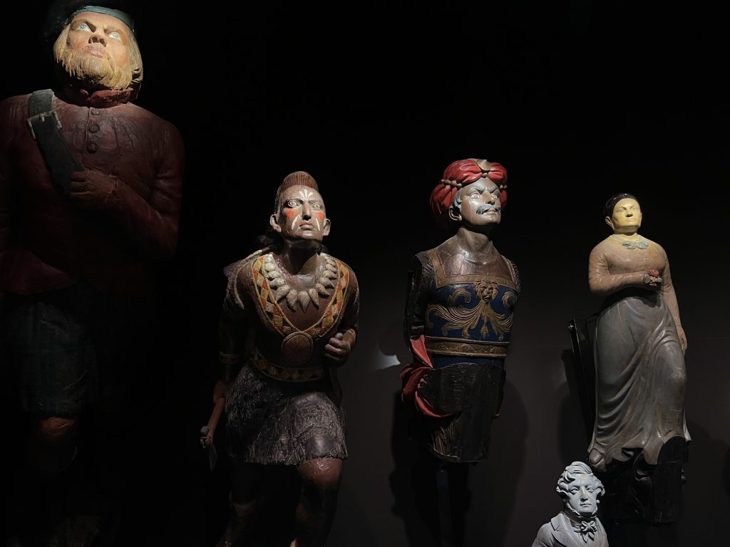 A black background filled with wooden figureheads from ships illuminated by up lighting so their faces are in bright light. There are 5 figureheads of different periods and cultures