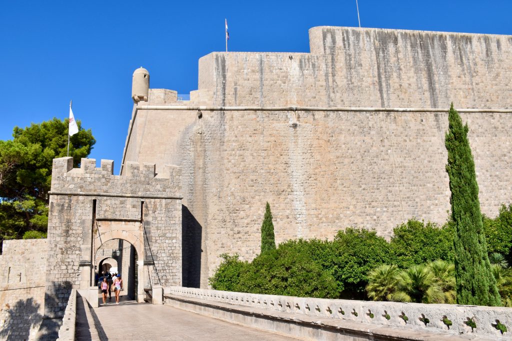 Looking along a stone bridge walkway to a castle gate with wooden drawbridge. To the right is a very tall stone fortress wall. Some trees either side. Blue sky above