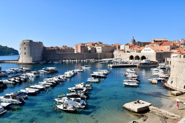 Looking down across a small port filled with rows of moored small white boats.To the right are the terracotta roofs of buildings within city walls. On the horizon are the walls of an old city with fortress at the left. Blue sky above. Turquoise waters of the old port