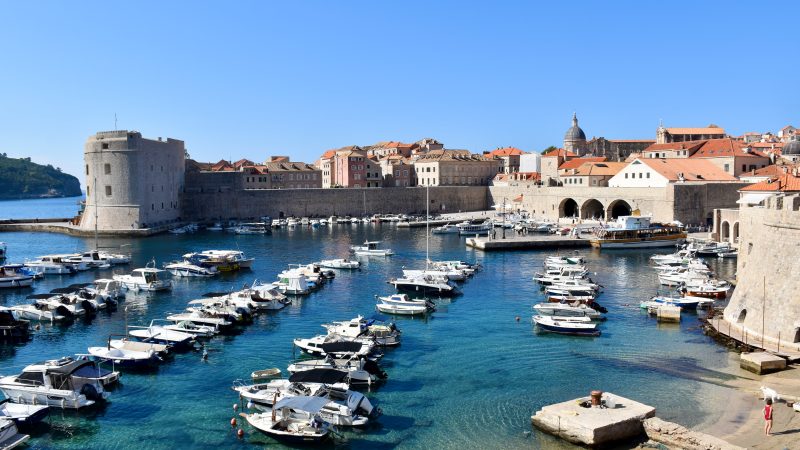 Looking down across a small port filled with rows of moored small white boats.To the right are the terracotta roofs of buildings within city walls. On the horizon are the walls of an old city with fortress at the left. Blue sky above. Turquoise waters of the old port