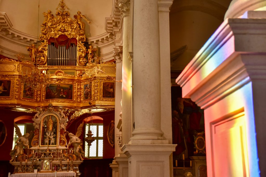 Inside a church. An ornate gold alter and organ appears to the left, decorated with old fashioned paintings. White columns to the middle and a white column to the right with rainbow colours reflected from something outside of the frame