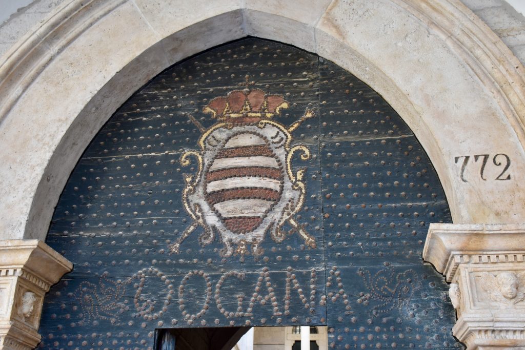 View of a stone archway over an old wooden door frame. A shield is drawn on the doorframe and the word 'Dogana' is studded into the woodwork. Studs decorate the rest of the frame.