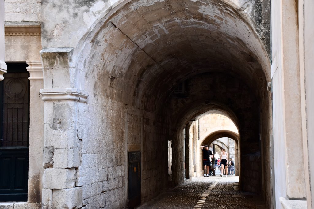 View through a long stone archway with cobbled pavement underneath. A couple in shorts and t-shirts are walking towards the camera in the distance