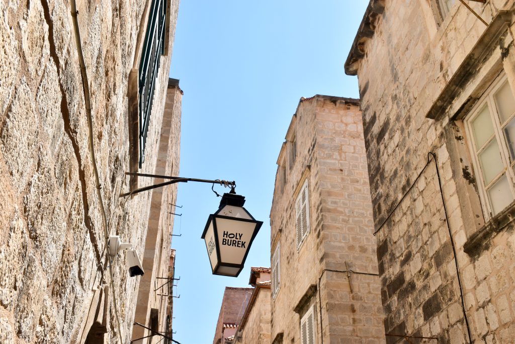 Looking up through the white stone walls of an alleyway towards a pale blue sky above. Tn the middle of the frame is a black iron lantern advertising 'Holy Burek' on the frosted glass