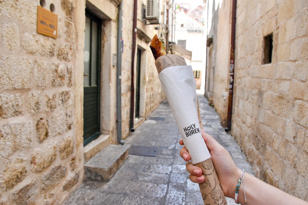 A hand with woven bracelet is holding a long thin sandwich up in the middle of the frame. It is wrapped in brown paper with a white napkin around the outside with the logo 'Holy Burek'. Alleyway behind