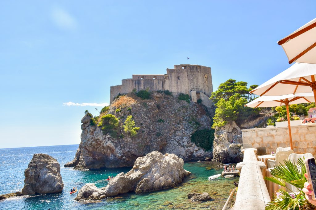View of a tall crop of stone with an old Fortress building on top. Turquoise see in front and to the left. A restaurant patio with cream sun parasols to the right.