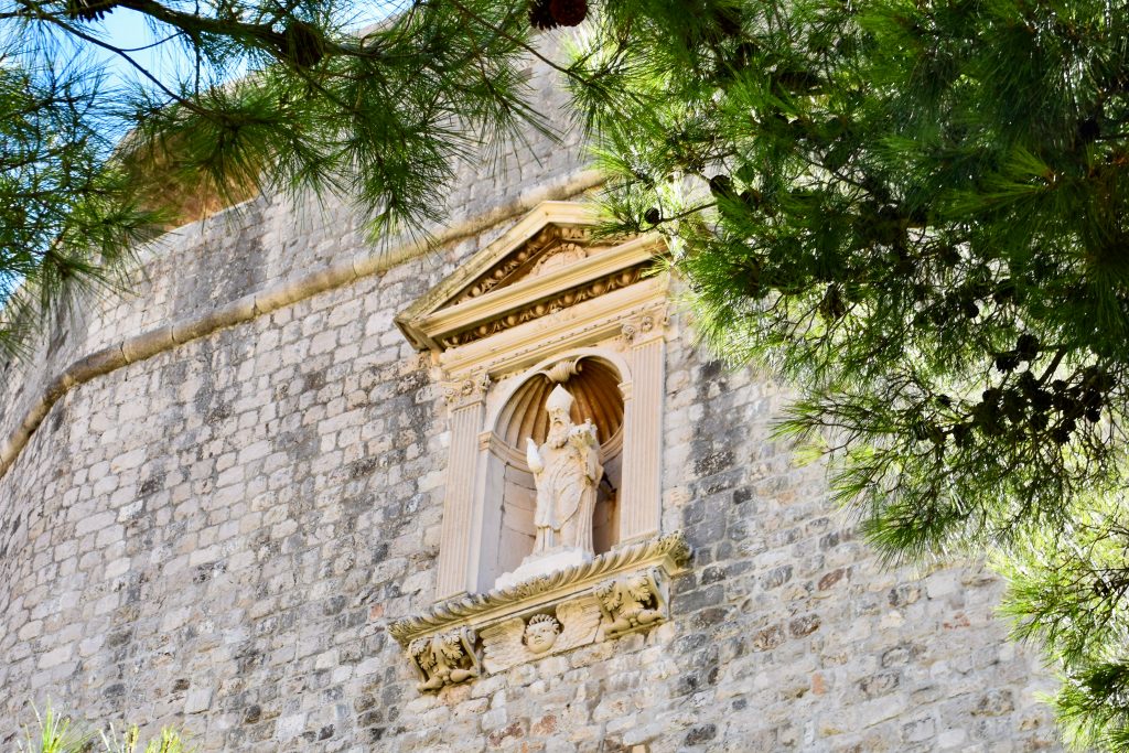 Looking up at tall stone wall with the lines of pine trees around the right and top. A stone statue of a priest or saint sits on an alcove in the middle of the wall.