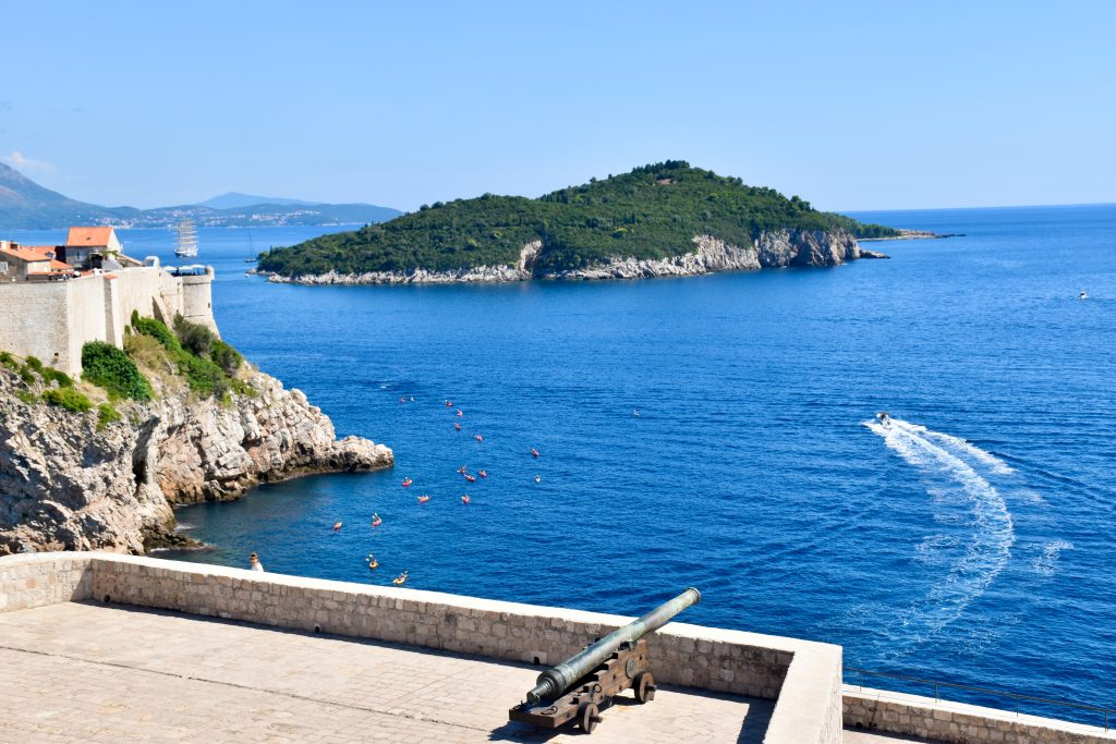 View from a stone fortress patio with cannon facing the sea. Walled fortresses to the left. Across the calm sea is a small rocky island covered in trees. Hills in the distance to the left. Pale blue sky above