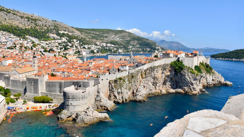 A walled city sits on rocky cliffs from the left hand side to the middle of the frame. The centre of the Old Town is crammed with tiled orange roofed buildings and some church towers. Hills behind. Water around the right hand side and a small pebble beach filled with orange kayaks to the bottom left